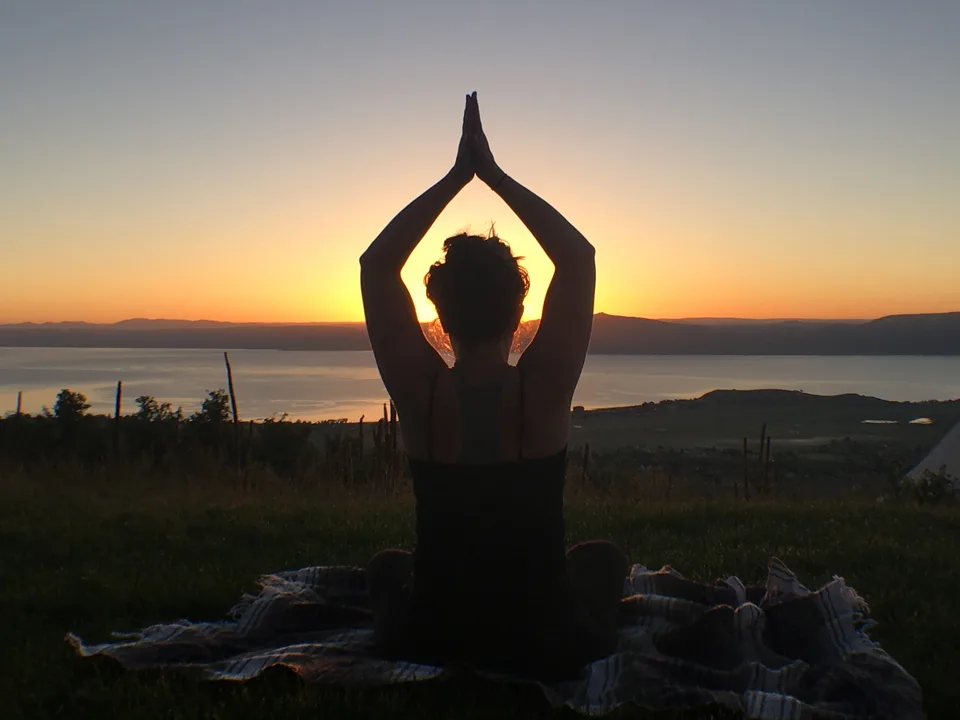 A woman practicing yoga beside the water at sunset.